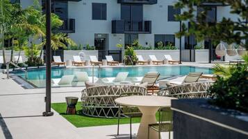 Zona de piscina moderna en un hotel, con sillones y mesas, rodeada por un edificio con balcones.