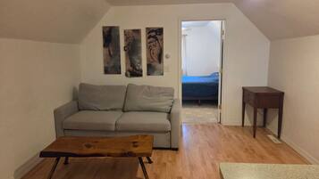 A living room with a grey sofa, a wooden coffee table, and a small wooden side table. There are abstract paintings on the wall and a door leading to another room.