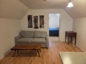 Een woonkamer met een grijze bank, een houten salontafel en een klein houten bijzettafeltje. Aan de muur hangen abstracte schilderijen en er is een deur naar een andere kamer.