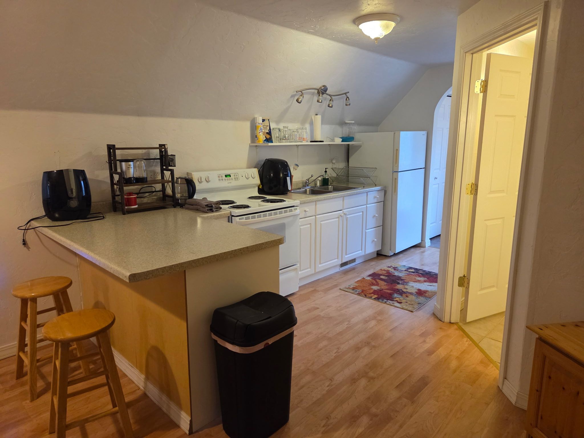 A kitchen with a central island, wooden stools, a trash bin, and a refrigerator.