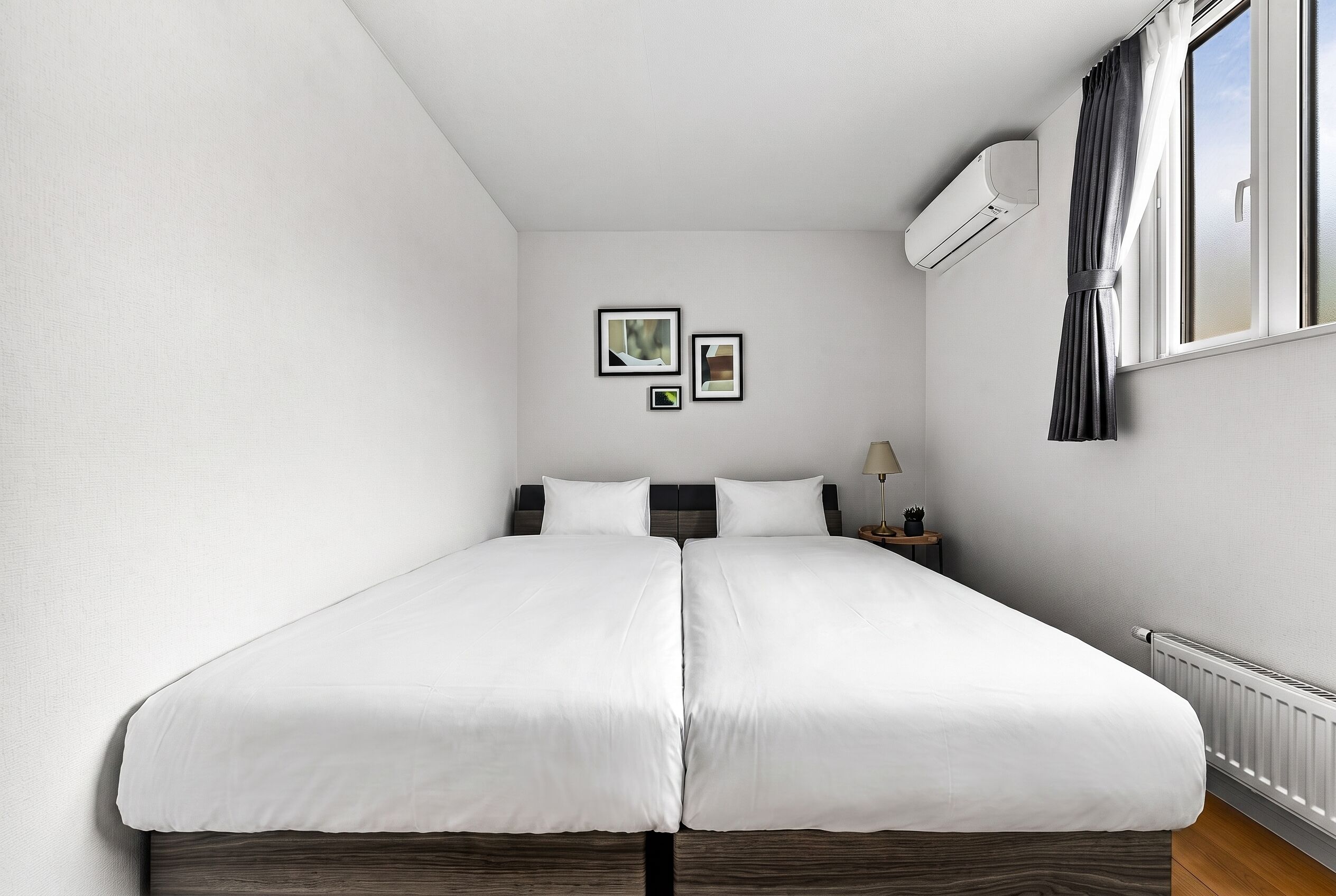 Una cama doble con ropa de cama blanca, una mesita de noche con una lámpara, y dos cuadros en la pared.