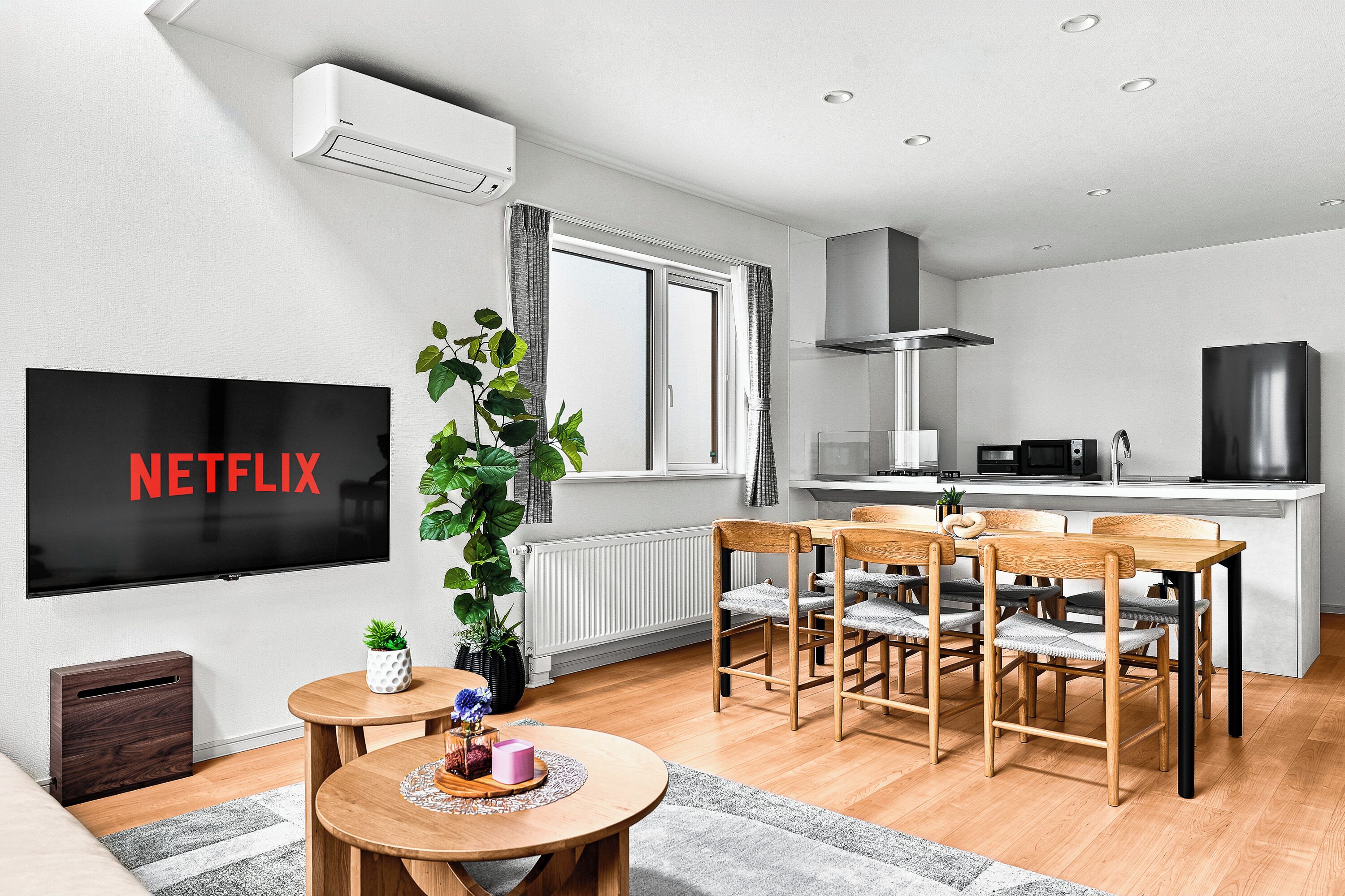 A modern living room with a flat-screen TV, a dining area with wooden chairs, a kitchen with a microwave, and a large window.