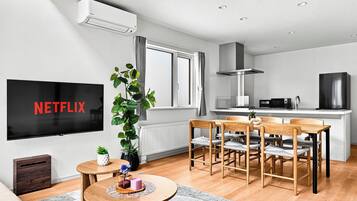 A modern living room with a flat-screen TV, a dining area with wooden chairs, a kitchen with a microwave, and a large window.