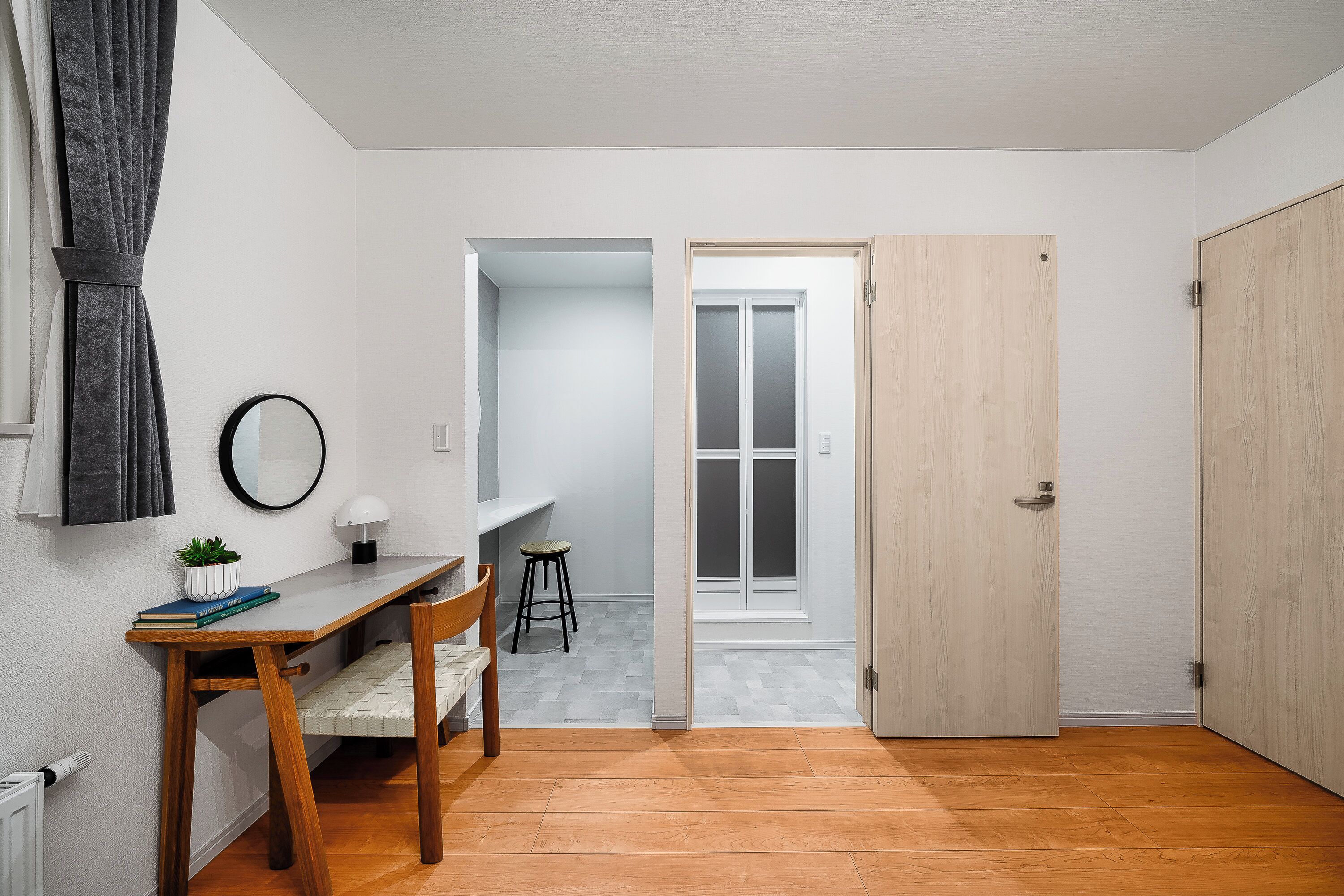 A room with a wooden floor, a desk with a chair, a small stool, a potted plant, and a door leading to another room.