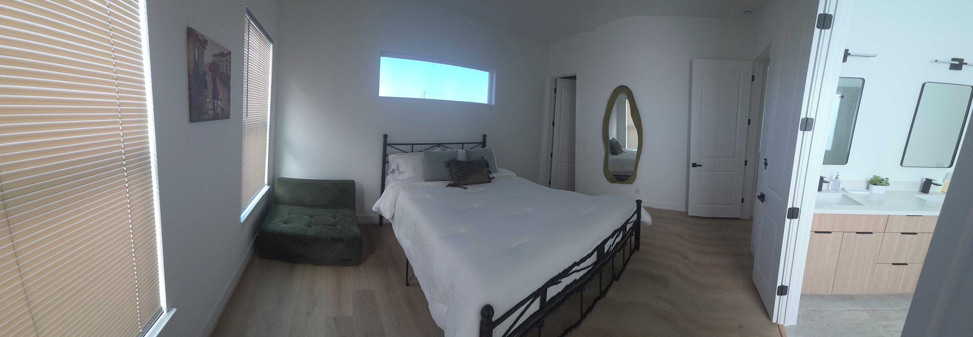 Une chambre à coucher avec un lit, un fauteuil vert, une fenêtre, une porte et une salle de bain avec un lavabo et un miroir.