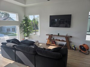 A modern living room with a sectional sofa, a flat-screen TV mounted on the wall, a wooden TV stand with books and decorative items, a potted plant, and a large window offering a view of the outside. - NEW! Houston Retreat, Near Medical District, NRG Stadium & Downtown (Houston)