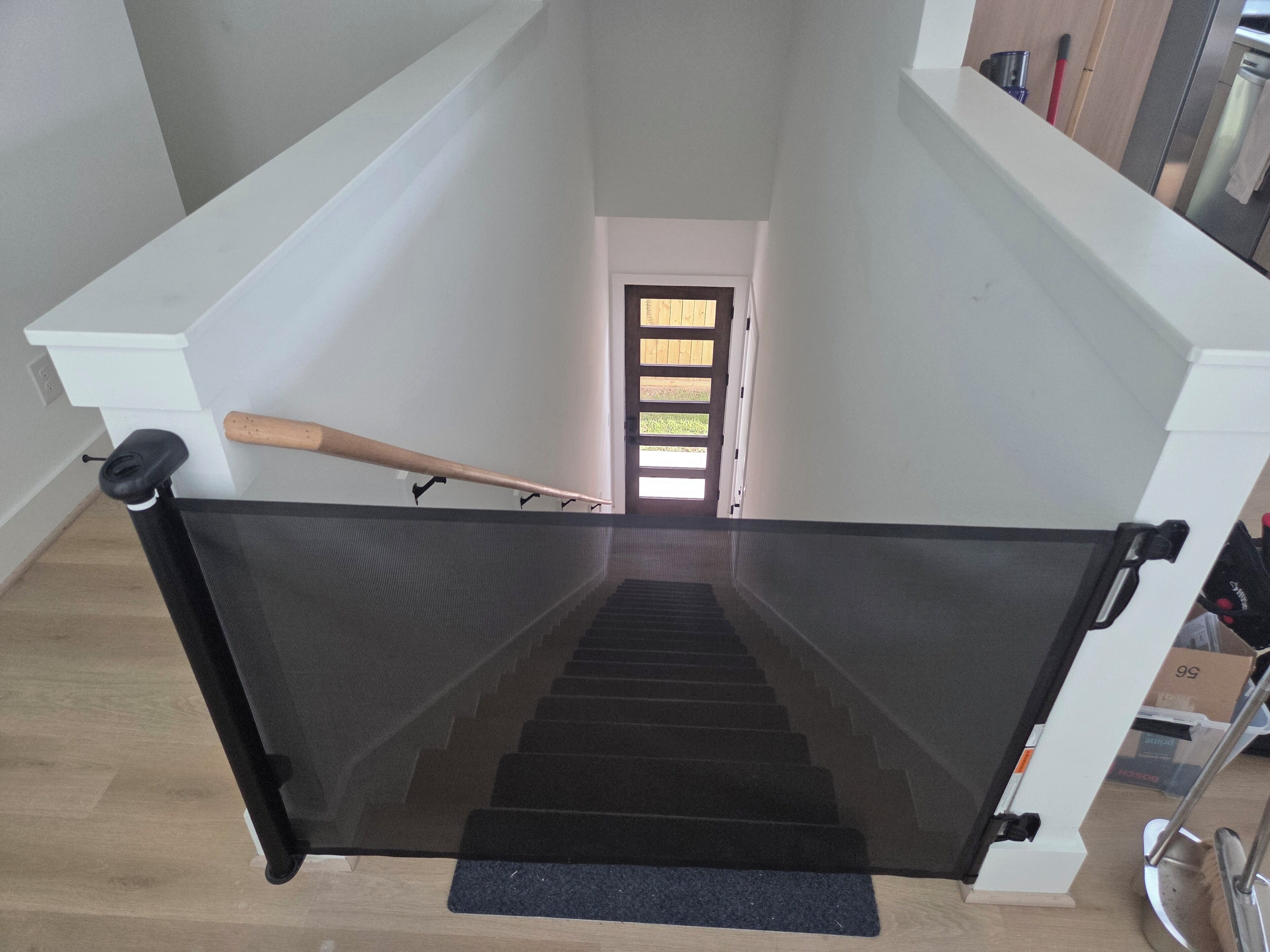 A staircase with a black safety gate, a wooden handrail, and a black mat at the bottom.