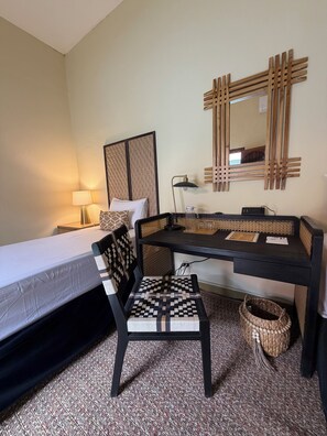 Ein Hotelzimmer mit einem Bett, einem Schreibtisch, einem Stuhl, einer Lampe, einem Spiegel und einem Korbwägelchen.