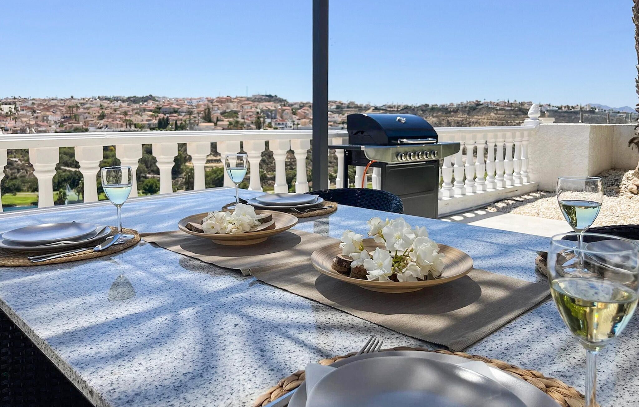 Un'area pranzo all'aperto con un tavolo imbandito per due, una griglia e una vista panoramica su una cittadina collinare.
