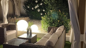 Un sofá de mimbre con cojines, una mesita y una planta en maceta, todo en un jardín con muro de piedra y plantas florales.