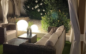 A wicker sofa with cushions, a small table, and a potted plant in a garden setting with a stone wall and flowering plants. - Lovely home in Rojales with sauna (Rojales)