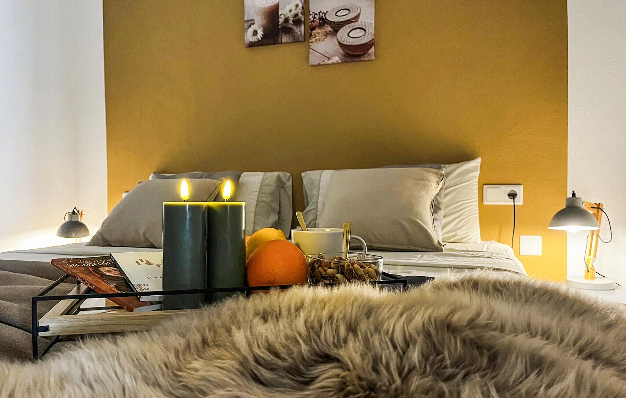 A bedroom with a bed, two lit candles, a cup, a book, and a tray with fruits.