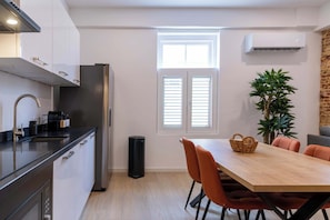 Eine moderne Küche mit einem Edelstahl-Kühlschrank, einer Spüle und einer Kaffeemaschine. Ein Esstisch mit Stühlen und einer Topfpflanze sind ebenfalls vorhanden.
