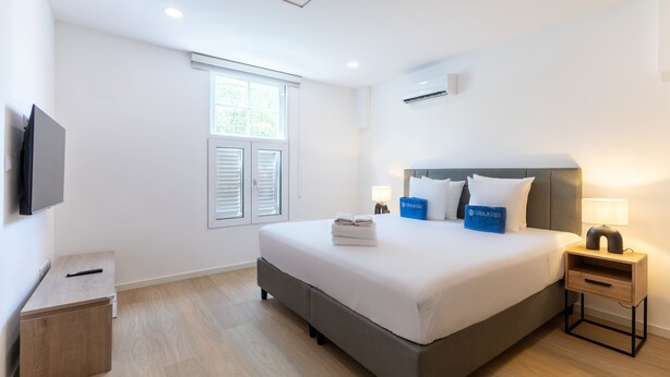Ein Hotelzimmer mit Bett, Nachttischen, einem Fernseher, einem Fenster mit Rollläden und einer Klimaanlage.