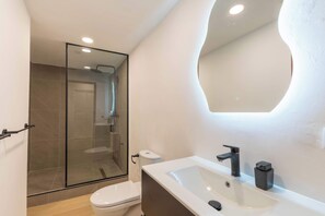 Un baño moderno con ducha envuelta en vidrio, inodoro blanco y lavamanos con espejo superior.