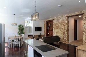 A modern kitchen with a central island, dining area with chairs, a hanging light fixture, and a brick accent wall. - NEW | Beautifully Restored Historic Apartment (Willemstad)
