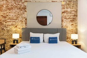 Habitación de hotel con pared de ladrillo, un espejo redondo sobre la cama, dos mesitas de noche con lámparas, una cama con ropa blanca y dos toallas azules con el logotipo del hotel.