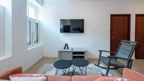 A modern living room with a flat-screen TV mounted on the wall, a grey entertainment unit, a round coffee table, a grey office chair, and a pink sofa. - Elegant Apartment in Historic Punda (Willemstad)