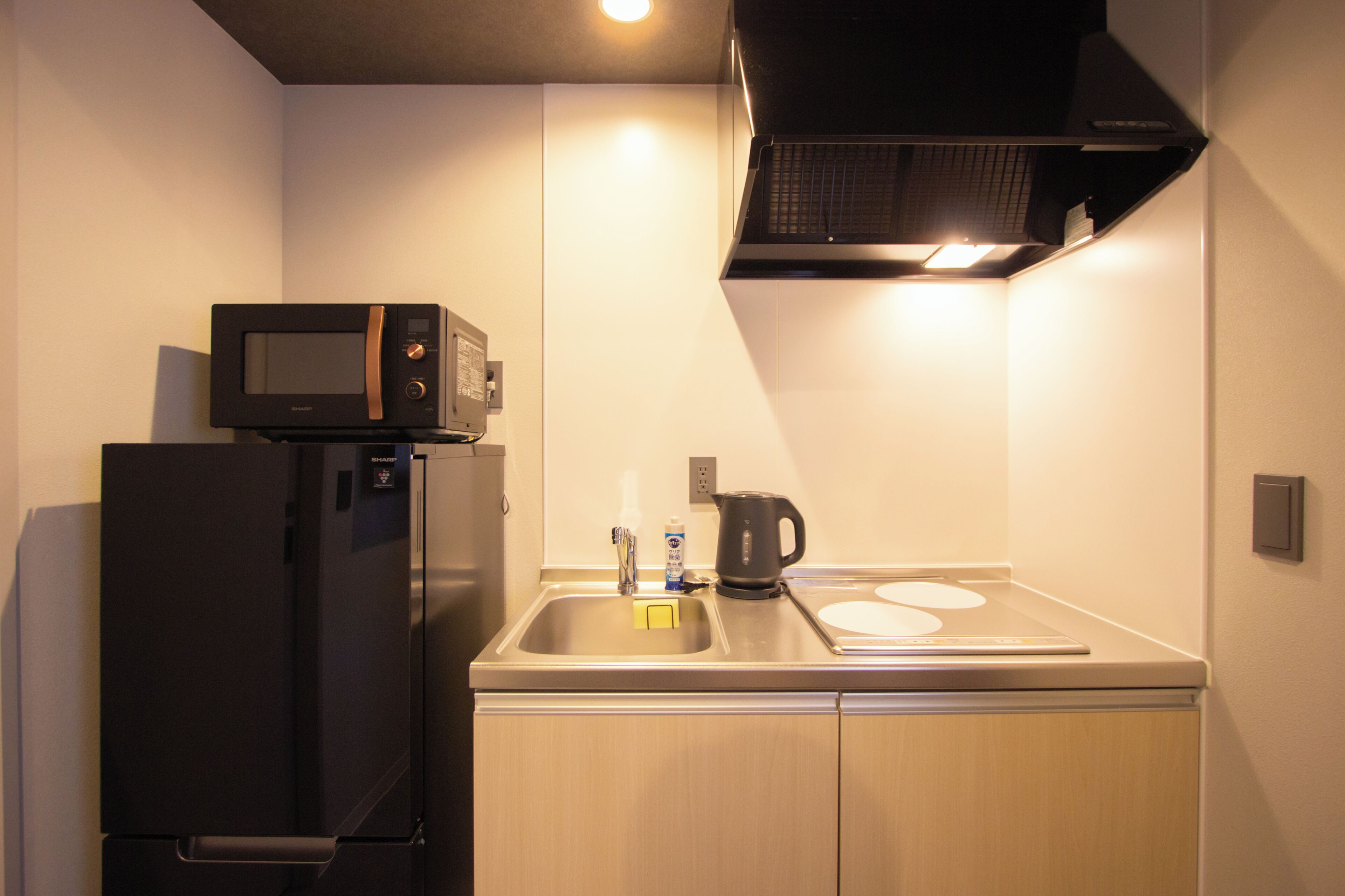 A compact kitchen with a microwave on top of a refrigerator, a kettle on the counter, and a sink with a faucet.