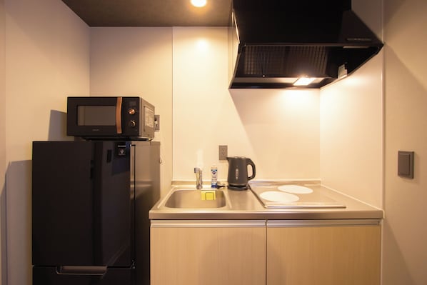 A compact kitchen with a microwave on top of a refrigerator, a kettle on the counter, and a sink with a faucet.