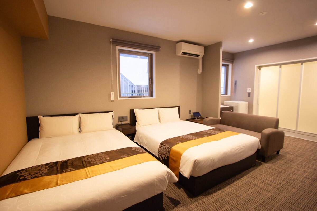 A hotel room with two beds, a sofa, a window, and an air conditioning unit.