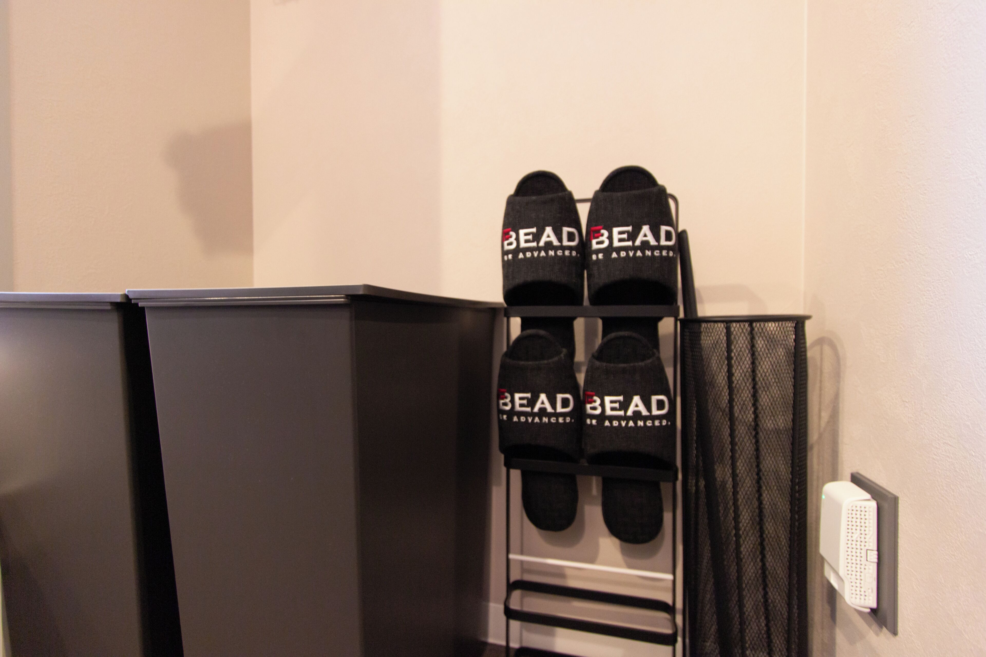 A pair of black slippers with 'BEAD BEAD' text on a rack next to a black cabinet and a white wall.