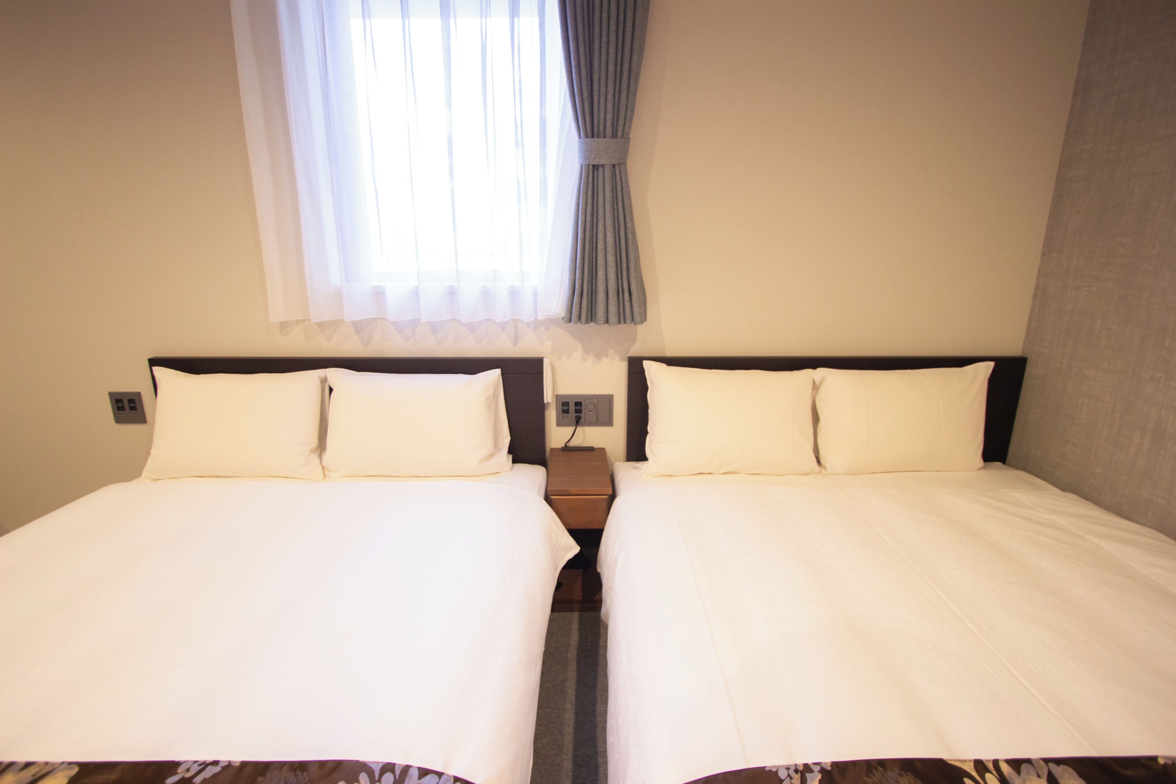 A double room with two beds, a window with sheer curtains, and a plain wall.