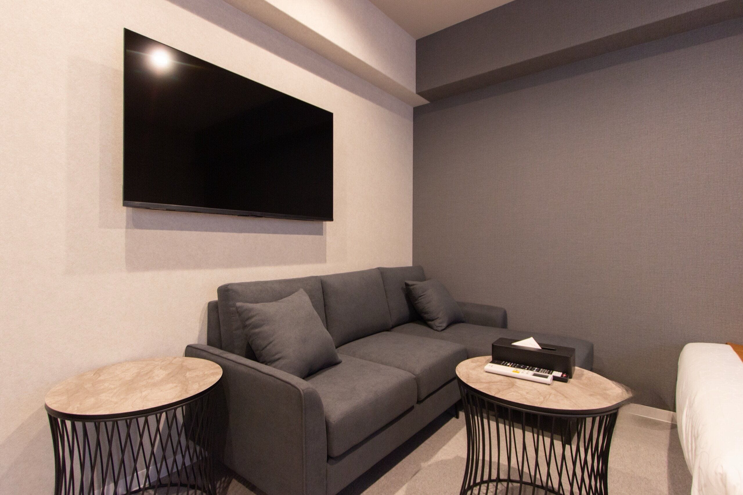 A modern living room with a grey sofa, two round side tables, a flat-screen TV mounted on the wall, and a small side table with a remote control.