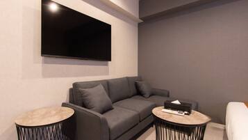 A modern living room with a grey sofa, two round side tables, a flat-screen TV mounted on the wall, and a small side table with a remote control.