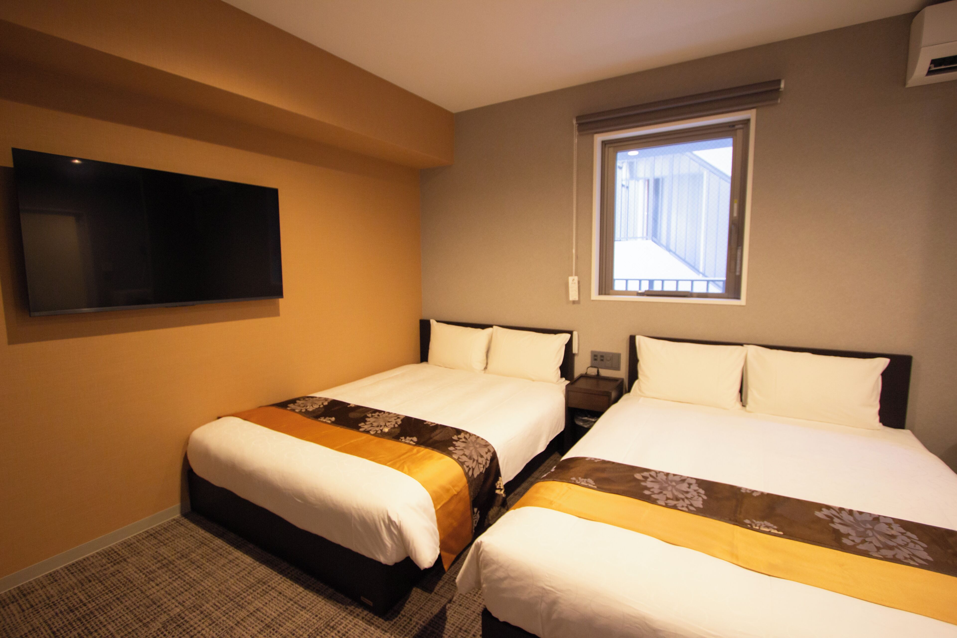 A hotel room with two beds, a flat-screen TV mounted on the wall, and a window providing a view of the outside.