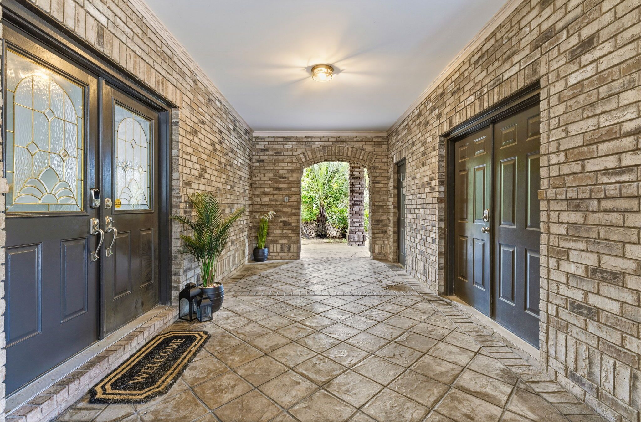 A brick-walled corridor with a tiled floor, a welcome mat, and potted plants.