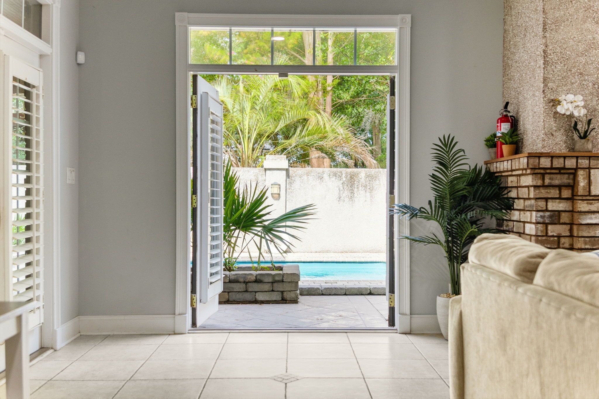 Una habitació amb una porta corredissa de vidre que dona a una zona de piscina, un espai d'seients amb coixins i una paret de maó amb plantes.
