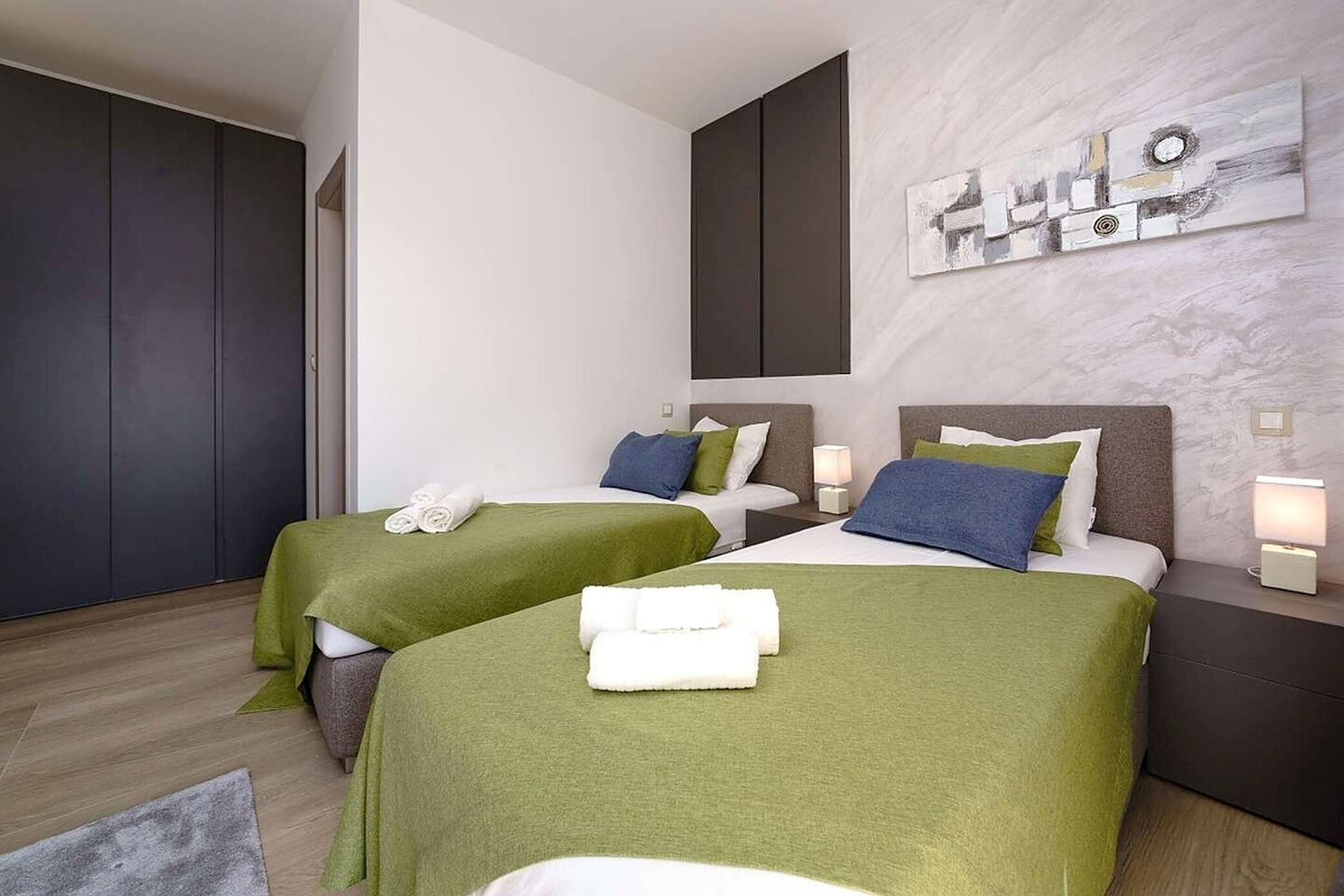 Ein Hotelzimmer mit zwei Betten, jedes mit einer grünen Tagesdecke und weißen Handtüchern. Darüber hängt ein großes, abstraktes Gemälde an der Wand.