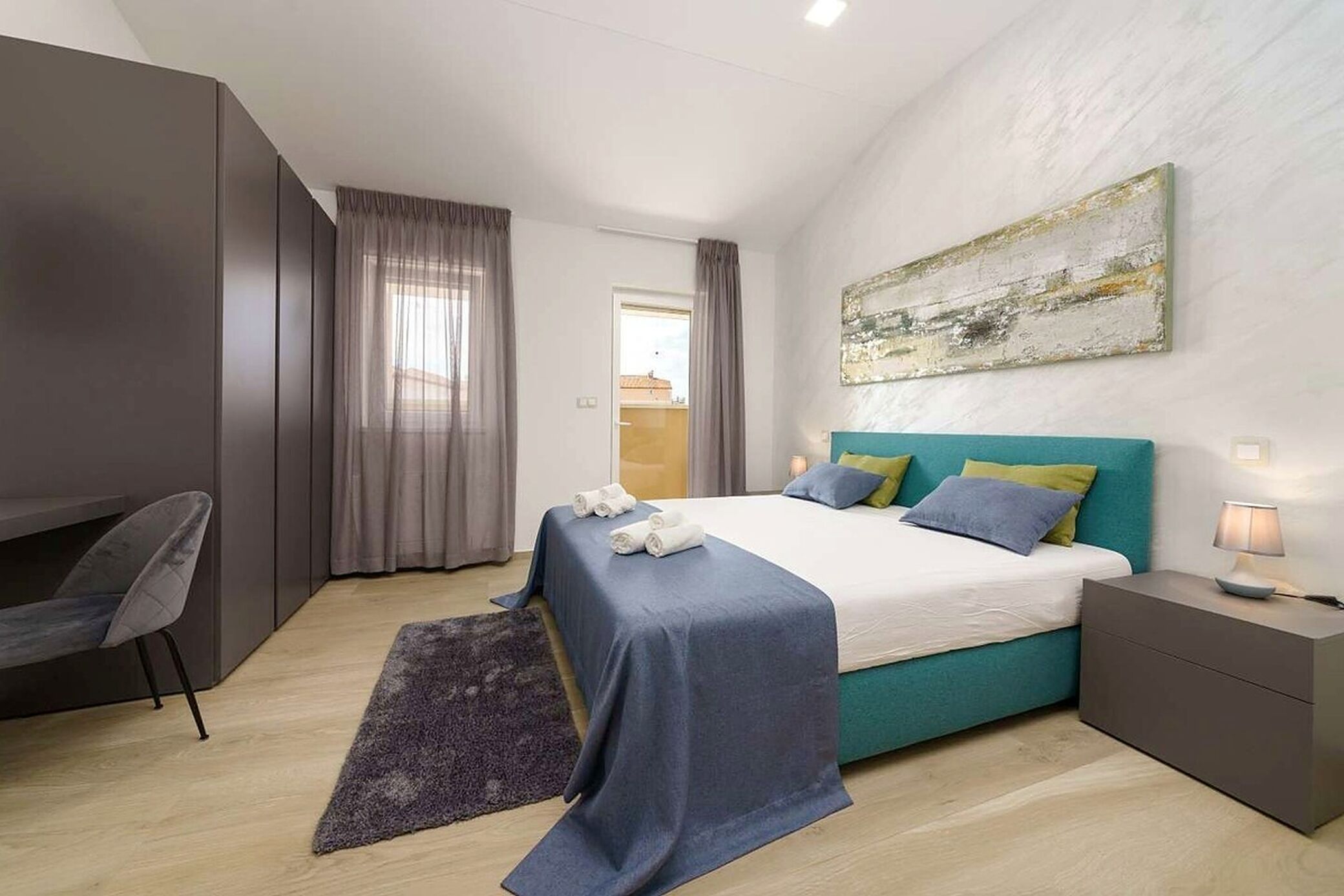 A modern hotel room with a bed, a desk, a chair, a nightstand, and a painting on the wall.