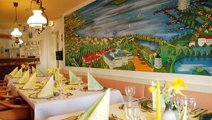 Un restaurant avec des tables dressées pour le service, une grande fresque murale et une vue sur la mer.