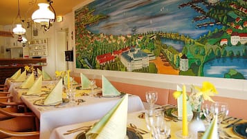 Een restaurant met gedekte tafels, een groot muurschilderij en hangende lampen.