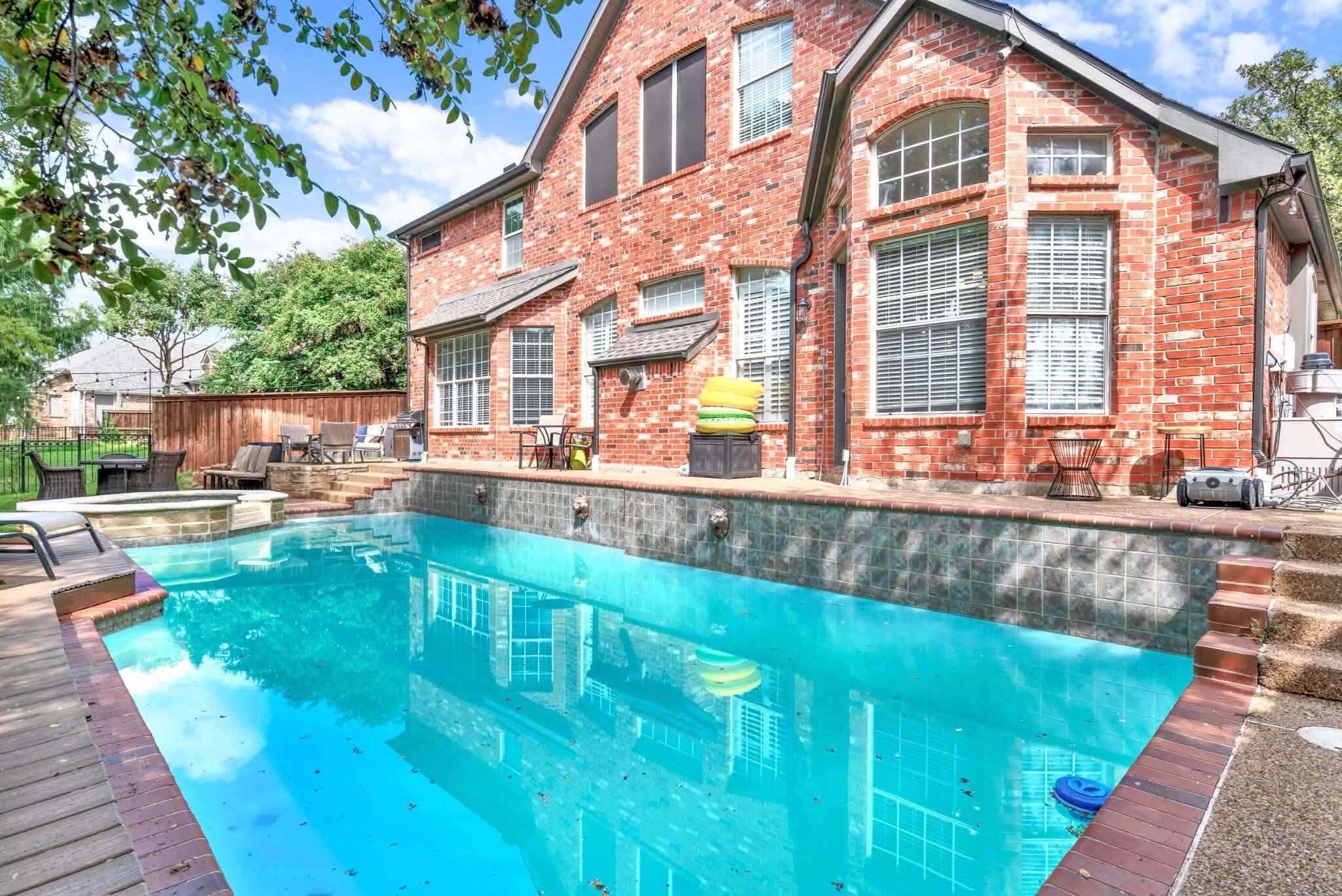 Une piscine avec une terrasse en bois, une maison en briques dotée de grandes fenêtres et des meubles d’extérieur.