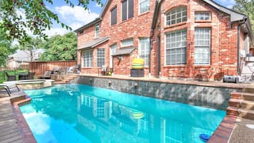 Une piscine avec une terrasse en bois, une maison en briques dotée de grandes fenêtres et des meubles d’extérieur.