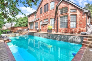 A swimming pool with a wooden deck, a brick house with large windows, and outdoor furniture.