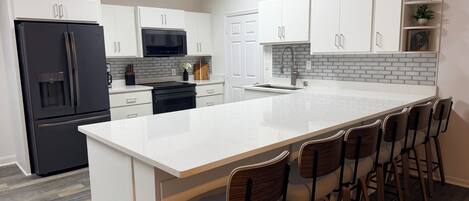 A modern kitchen with white cabinetry, a black refrigerator, a stainless steel range, and a bar area with stools.