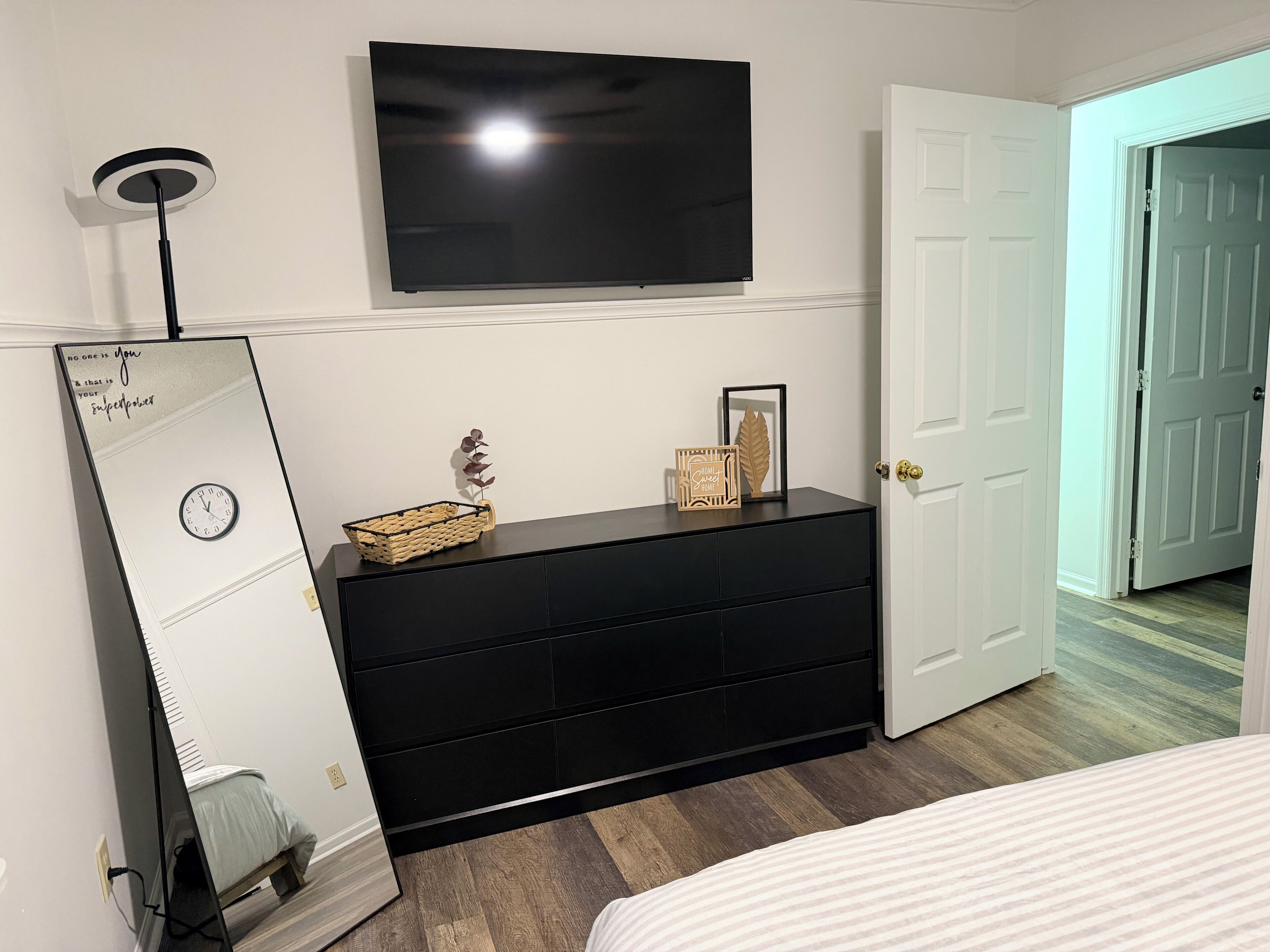 A bedroom with a bed, a black dresser, a television, a mirror, and a door.
