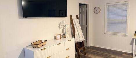 Une pièce dotée d’un grand téléviseur à écran plat fixé au mur, d’une commode blanche avec des tiroirs, d’un chevalet en bois recouvert d’un tissu blanc, d’une petite table blanche décorée d’un ananas et d’une fenêtre avec des stores.