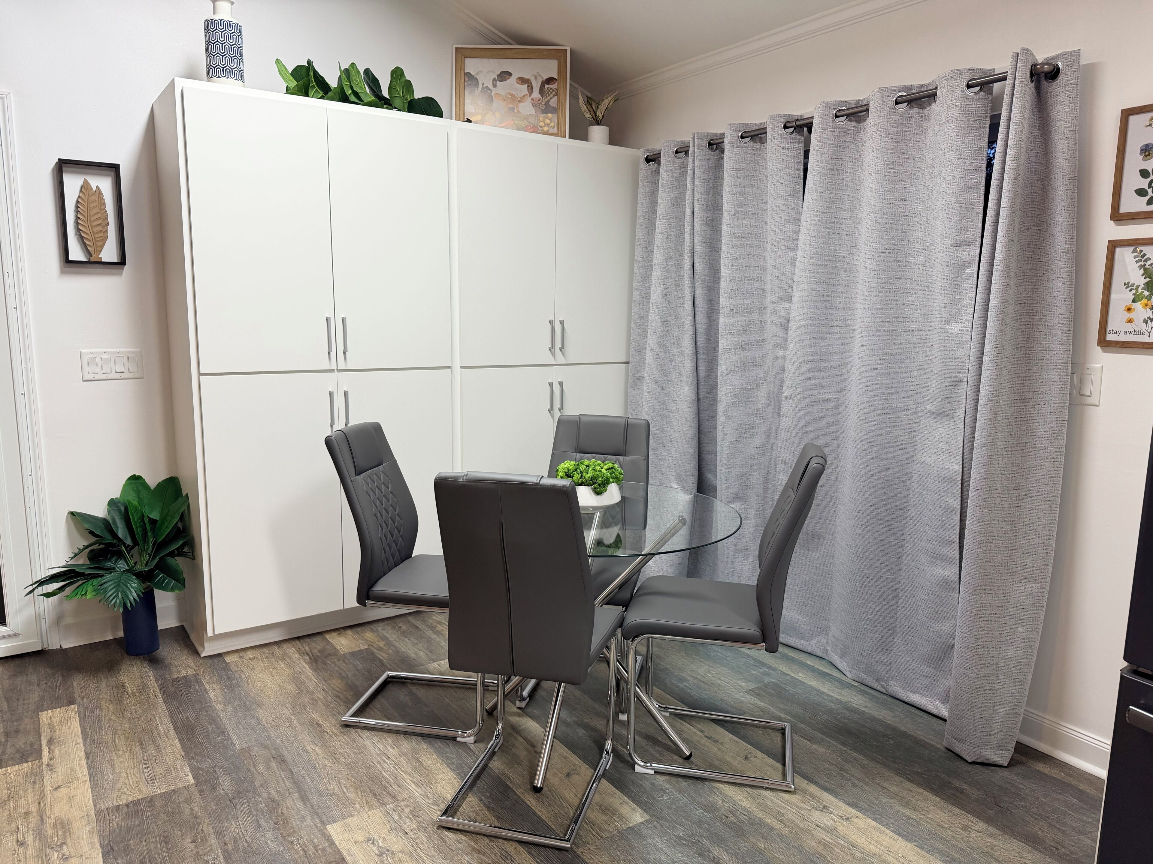 A modern dining area with a glass table, metal chairs, a white cabinet, and grey curtains.