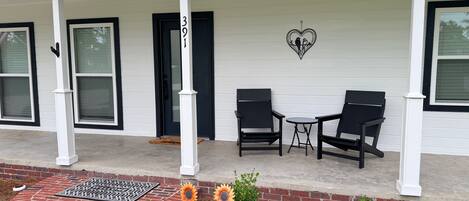 A porch with two chairs and a small table, a brick pathway leading to the door, and a heart-shaped decoration on the wall.