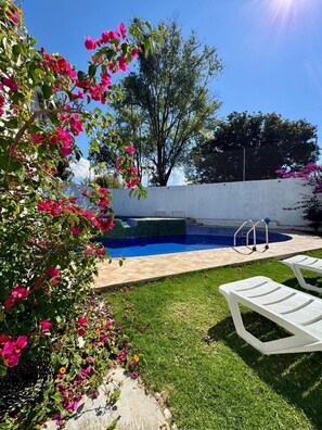 Una piscina con un cerco blanco, un árbol y una zona verde con sillas de descanso.
