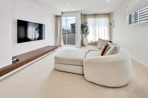 A modern living room with a large flat-screen TV mounted on the wall, a sectional sofa, a wooden coffee table, and a sliding glass door leading to a balcony. - Villa Laya, Palm Beach (Palm Beach)