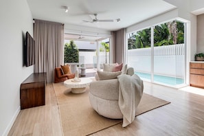 A modern living room with a large window, a flat-screen TV mounted on the wall, a round coffee table, and a sofa with a throw blanket. - Villa Laya, Palm Beach (Palm Beach)