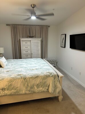 Une chambre à coucher avec un lit, un ventilateur de plafond, une commode, un téléviseur et une fenêtre avec des rideaux.