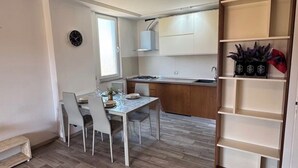 Een kleine keuken met witte kastjes, een eettafel met stoelen en een klein raam.