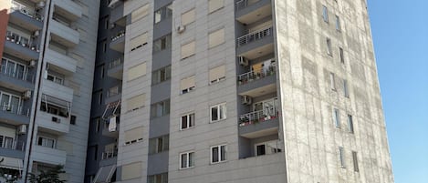 A multi-story residential building with balconies, parked cars, and a clear sky.